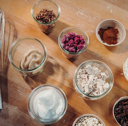 Mehrere kleine Glasschälchen auf einer Holzarbeitsfläche, gefüllt mit Backzutaten wie Zucker, Kakaopulver, Schokolade, getrockneten Beeren und Streuseln.
